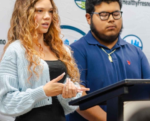 A young female participant in a light-blue cardigan presenting at the Fresno BHC Youth Internships Program podium alongside a male teammate, highlighting leadership development and encouraging more youth to apply.