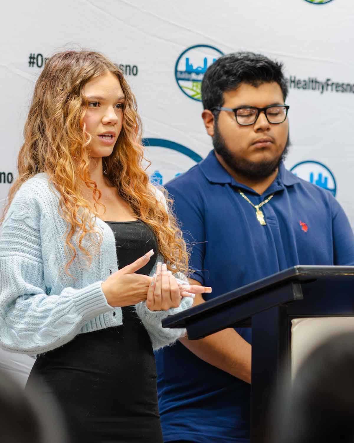 A young female participant in a light-blue cardigan presenting at the Fresno BHC Youth Internships Program podium alongside a male teammate, highlighting leadership development and encouraging more youth to apply.