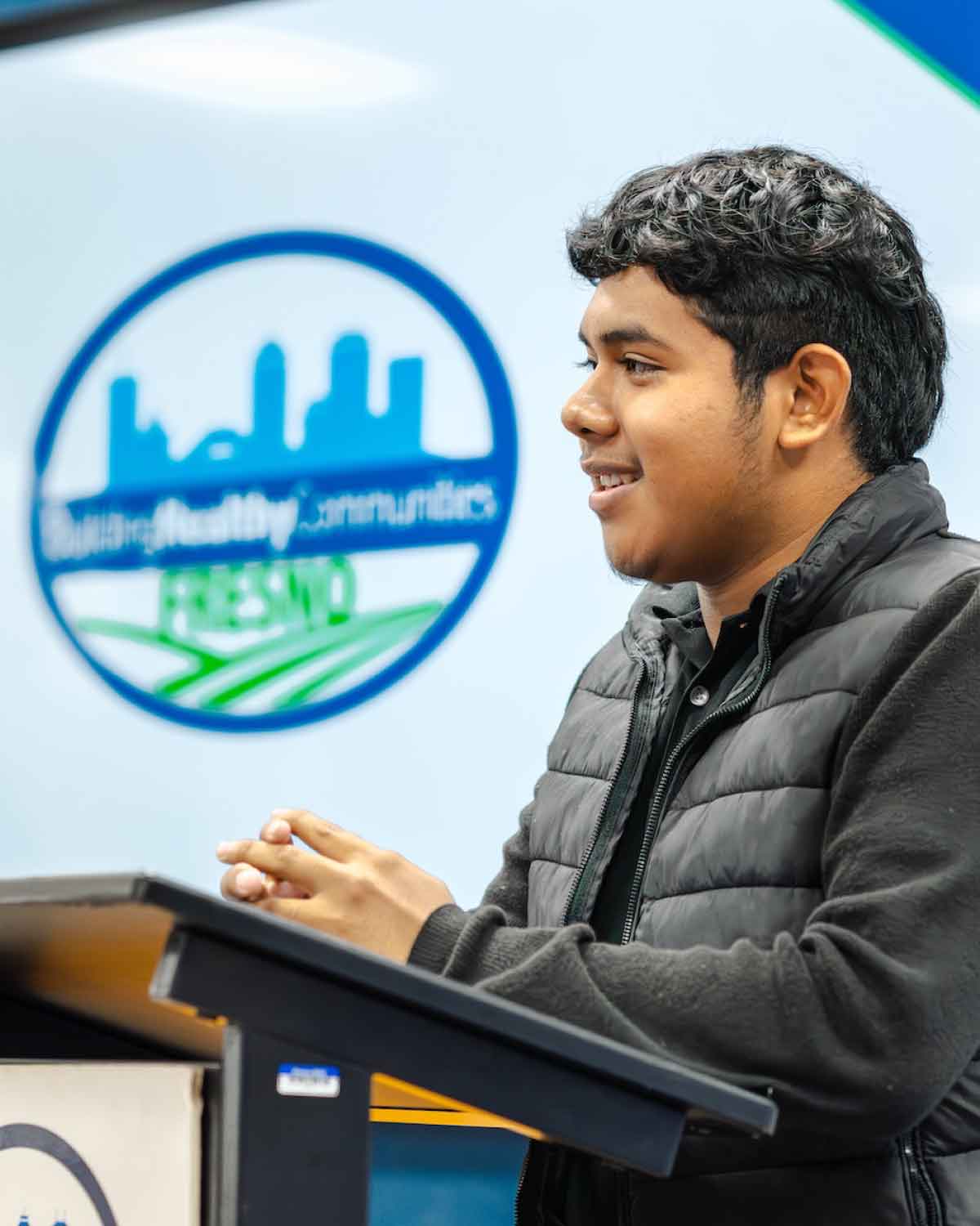 A motivated young man in a black vest speaking at a podium with the Building Healthy Communities Fresno logo, exemplifying the program’s emphasis on youth empowerment and leadership, and inspiring others to apply.