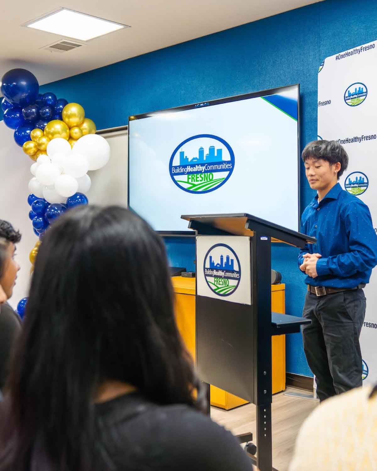 A youth presenter in a blue shirt speaking at a podium with the Building Healthy Communities Fresno logo displayed behind him, demonstrating leadership and encouraging peers to apply to the Youth Internships Program.