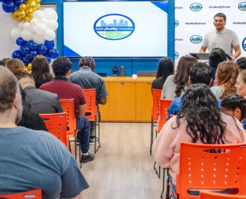 A wide shot of the Fresno BHC Youth Internships Program audience seated in bright orange chairs, watching a speaker in front of a large screen showing the Building Healthy Communities Fresno logo, emphasizing a supportive environment for youth empowerment.