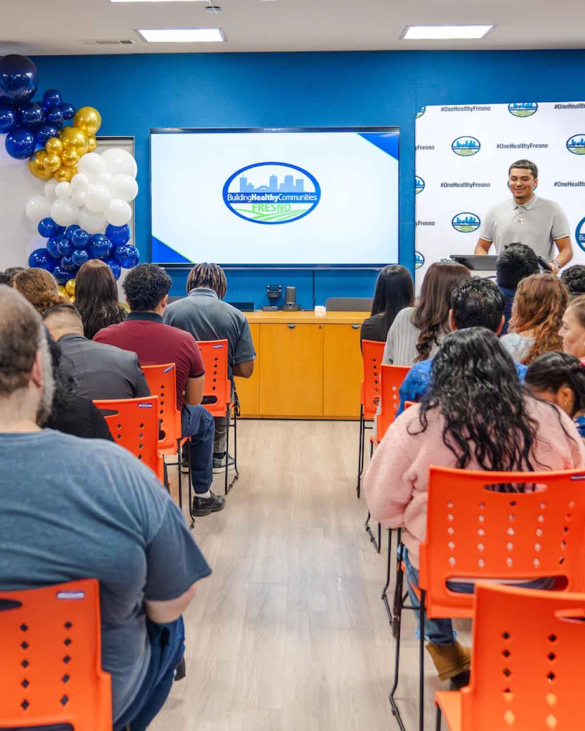 A wide shot of the Fresno BHC Youth Internships Program audience seated in bright orange chairs, watching a speaker in front of a large screen showing the Building Healthy Communities Fresno logo, emphasizing a supportive environment for youth empowerment.