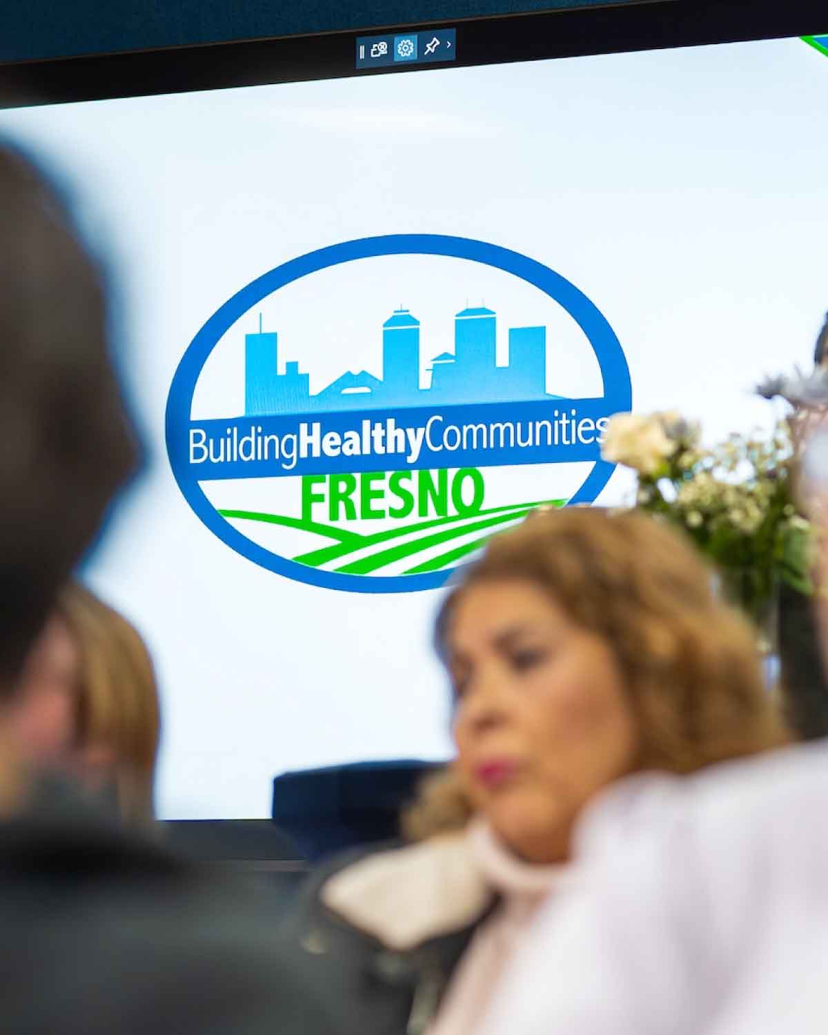 A close-up of the Building Healthy Communities Fresno logo displayed on a large screen, with attendees in the foreground, symbolizing the organization’s commitment to empowering youth through internships and advocacy.