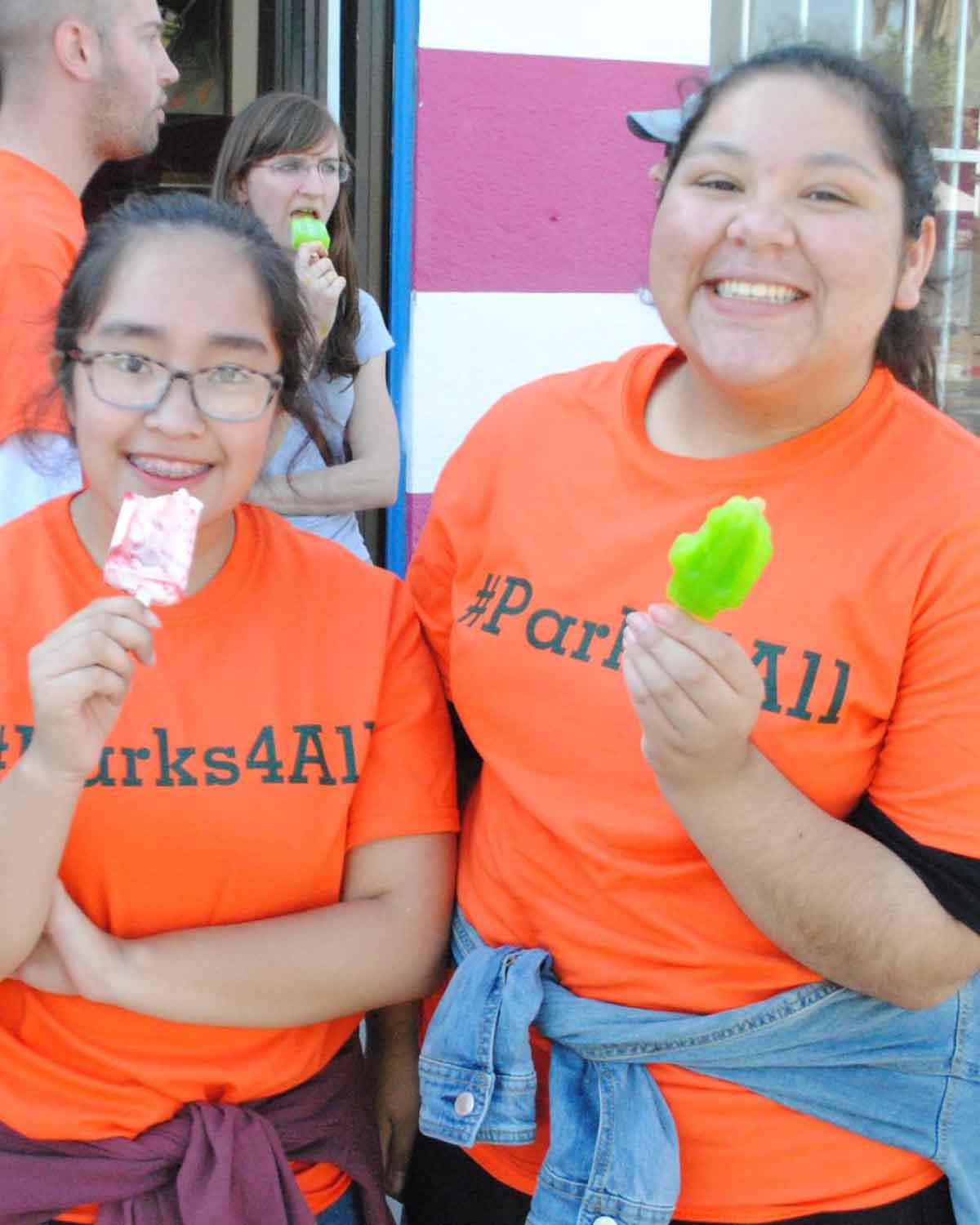 Two smiling teens wearing bright orange #Parks4All T-shirts and enjoying popsicles during a Fresno BHC community event, showcasing fun, inclusive engagement that inspires others to apply for youth leadership opportunities.