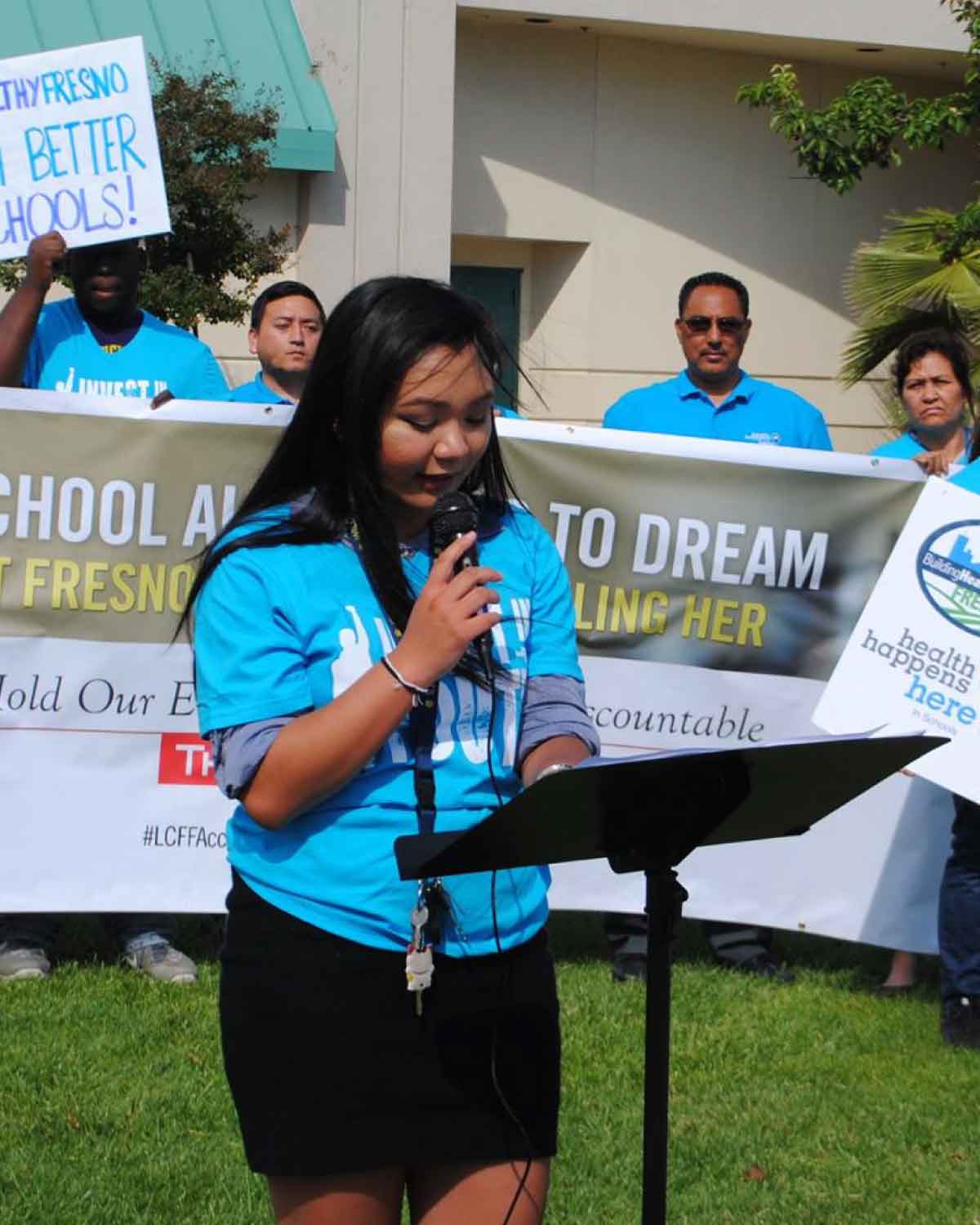 A young speaker in a blue ‘Invest in…’ T-shirt addressing the crowd at a Fresno BHC rally, passionately advocating for better schools and illustrating the power of youth leadership through the Youth Internships Program.