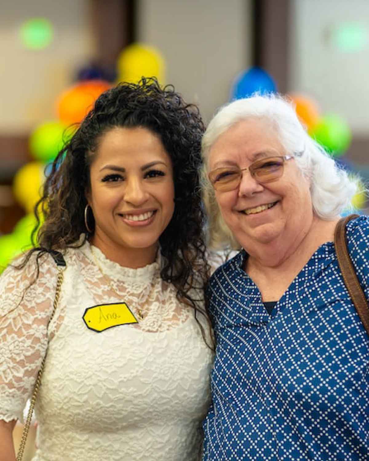 Two women standing close together and smiling at the camera, one in a white lace blouse with a name tag, and the other in a blue patterned top and glasses, at a community event with colorful balloons in the background.