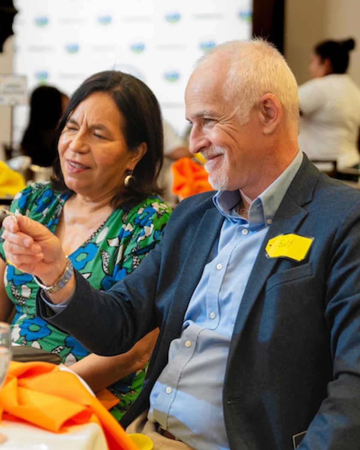 A man in a blazer and a woman in a green floral blouse seated at a table, both smiling and engaged in conversation during an event.