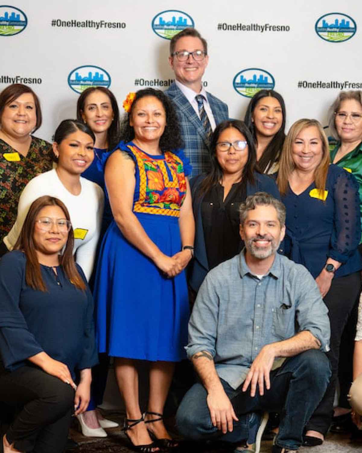 A group of people posing together in front of a step-and-repeat banner with ‘#OneHealthyFresno’ logos, wearing a mix of professional and traditional outfits and smiling for the camera.