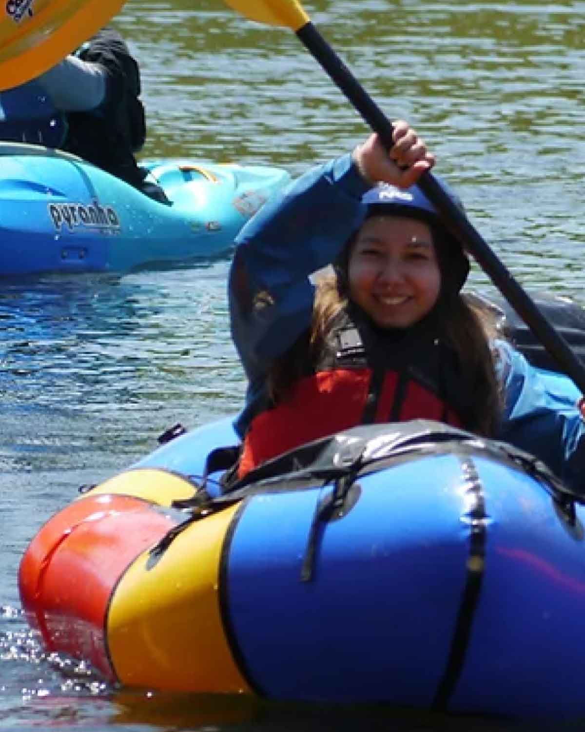 A person paddling a brightly colored inflatable kayak on calm water, smiling and holding a yellow paddle overhead.