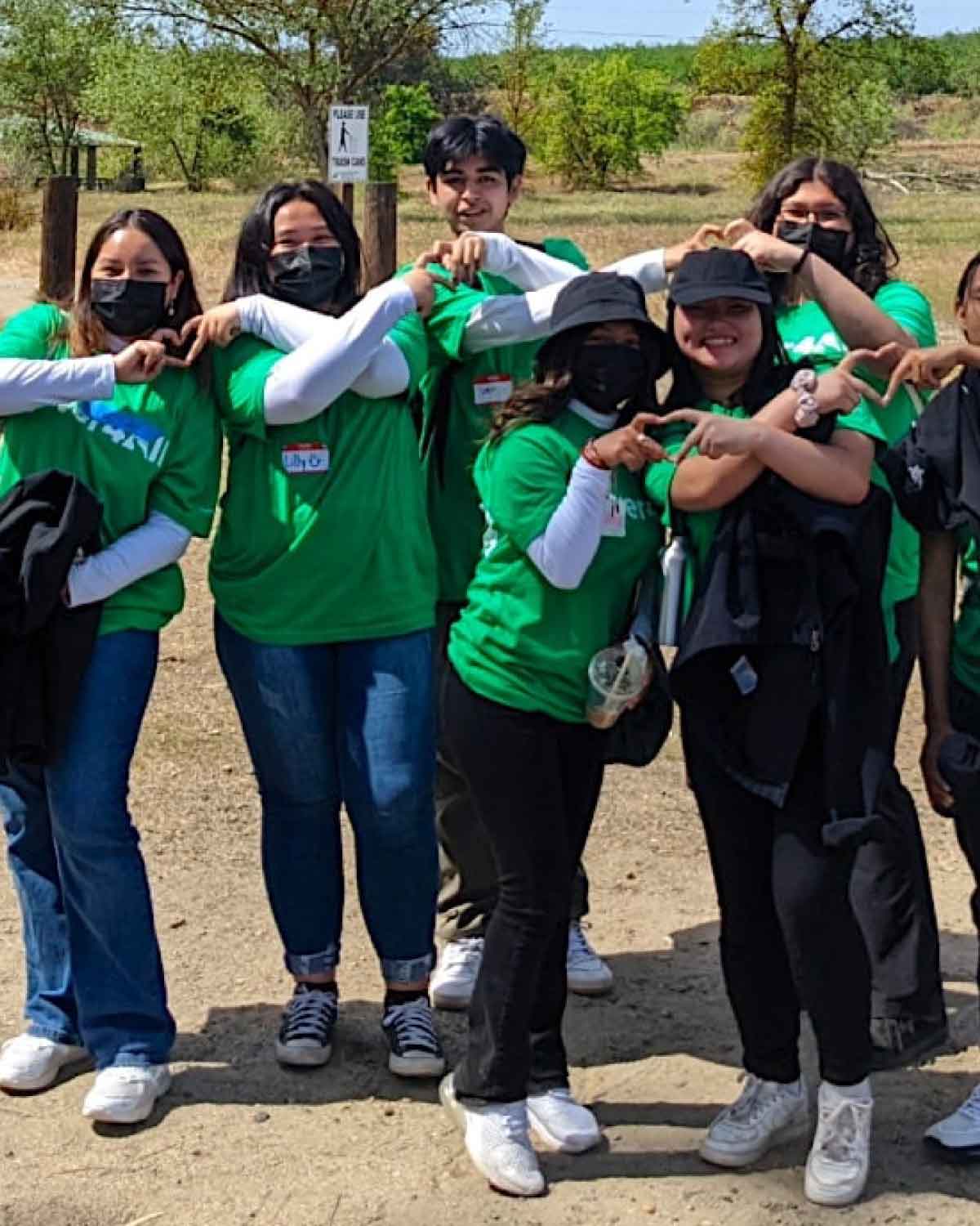 A group of young people in matching green T-shirts and masks standing outdoors, forming heart shapes with their hands.