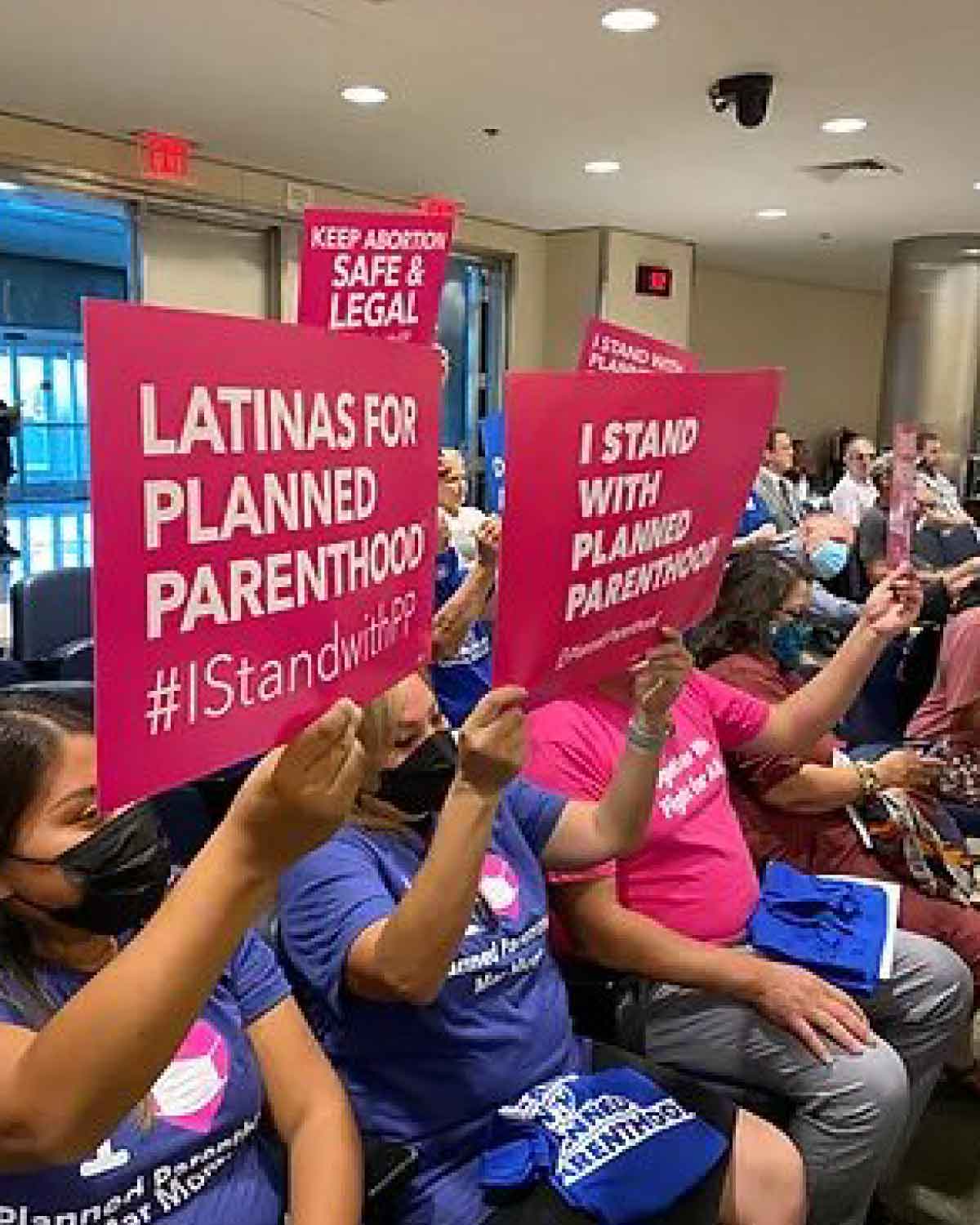 Supporters seated indoors holding pink signs that read ‘Latinas for Planned Parenthood,’ ‘Keep Abortion Safe & Legal,’ and ‘I Stand With Planned Parenthood.’