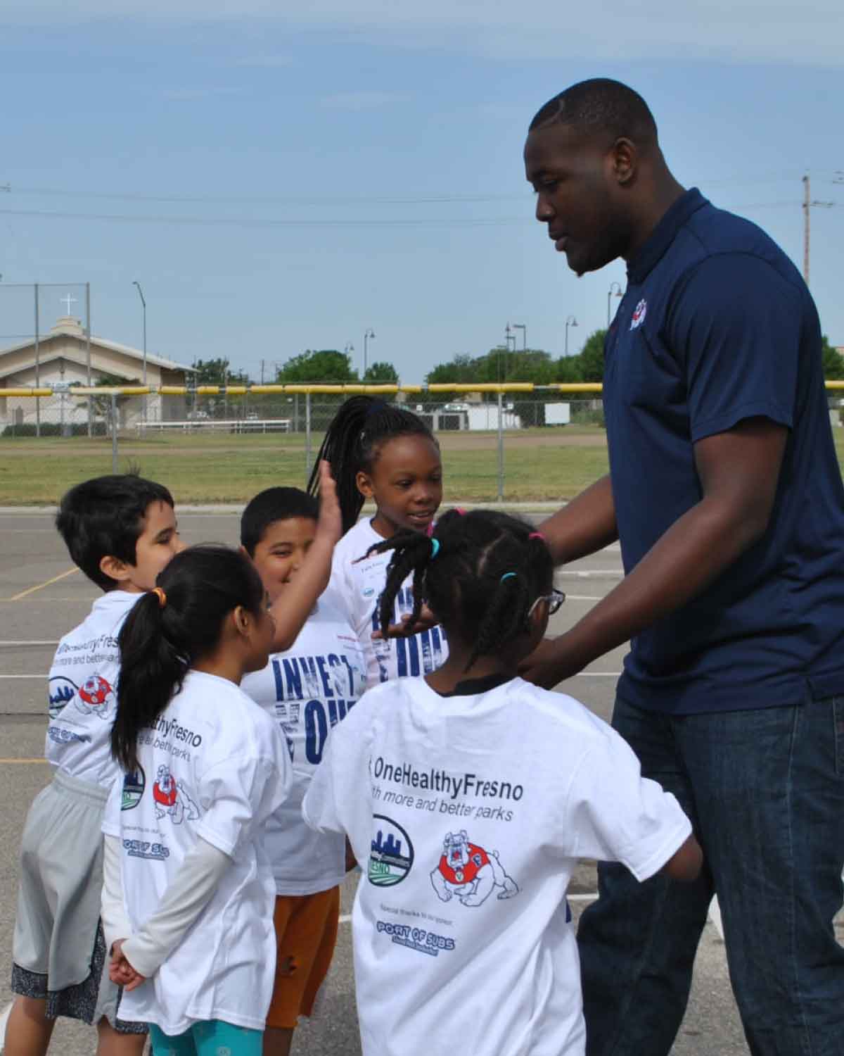 An adult in a navy polo shirt interacting with a group of children wearing white ‘#OneHealthyFresno’ T-shirts on a schoolyard.