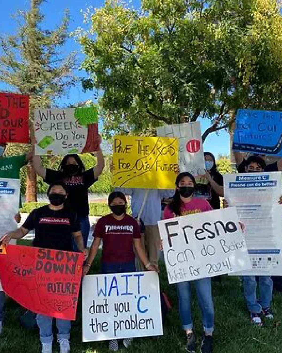 A group of masked individuals outside holding handmade signs with messages like ‘Fresno Can Do Better,’ ‘Wait, Don’t You ‘C’ the Problem,’ and other slogans advocating for change.