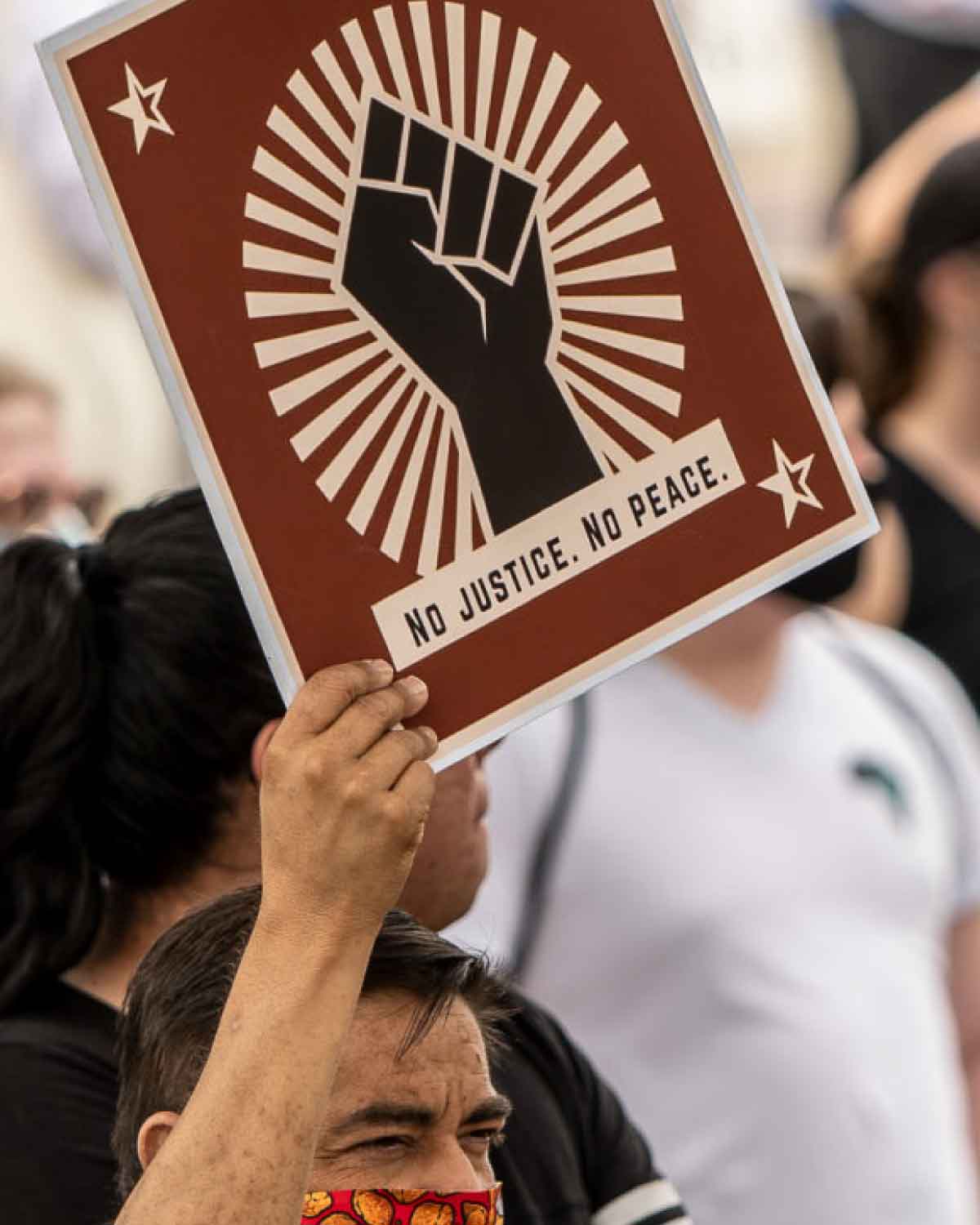 A protester in a crowd holding a large, rust-colored sign showing a raised fist surrounded by radiating lines and the text ‘No Justice. No Peace.’