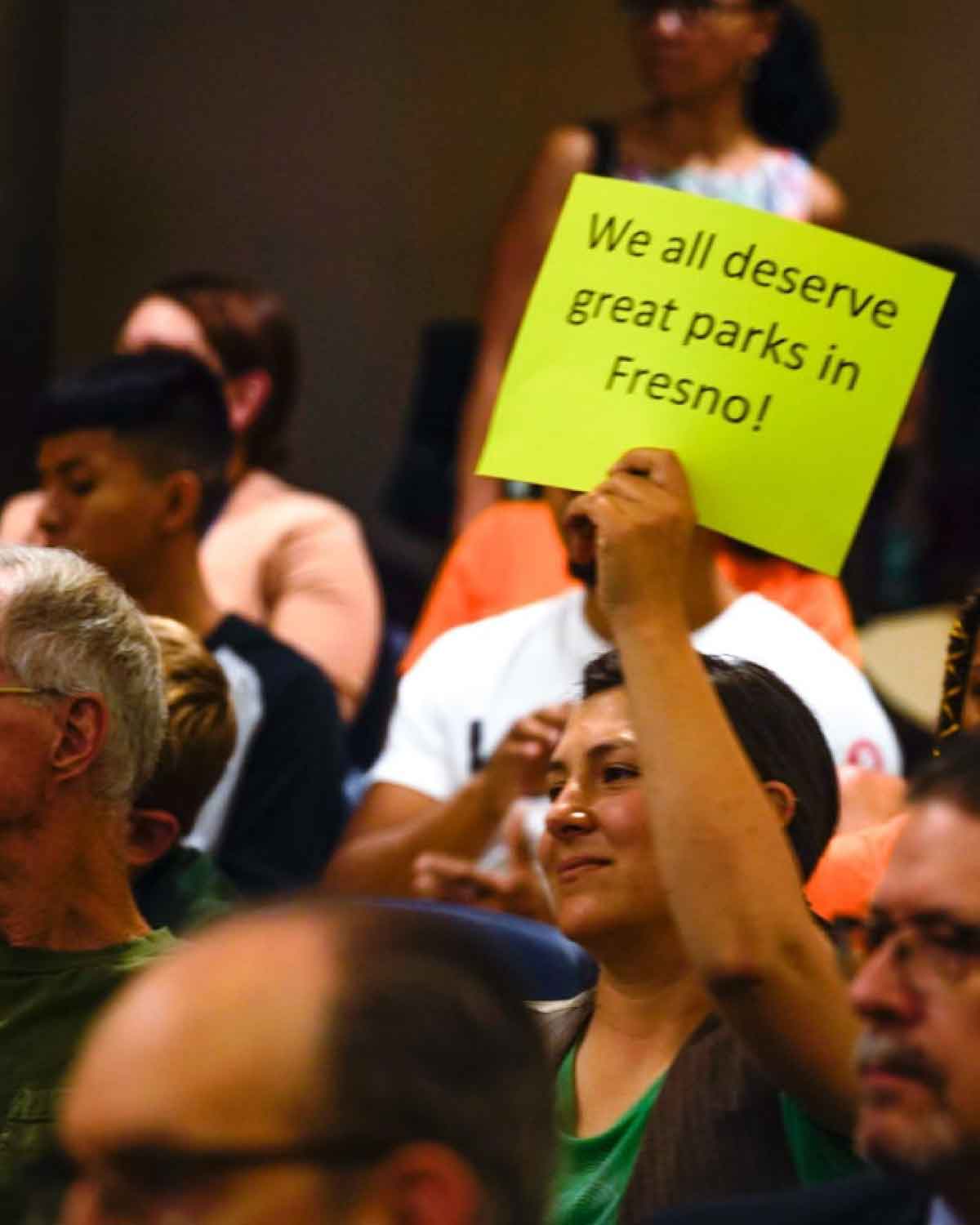 An attendee at a public meeting raising a neon-green sign that reads ‘We all deserve great parks in Fresno!’ while others look on.