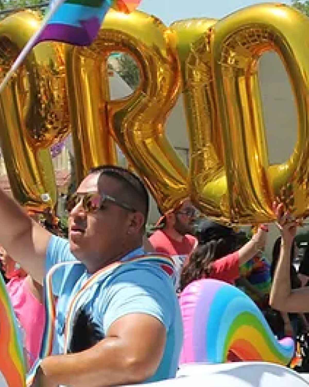 A participant in a Pride parade waving a rainbow flag and marching beside large gold balloon letters spelling ‘PRIDE,’ surrounded by other colorful floats and marchers.