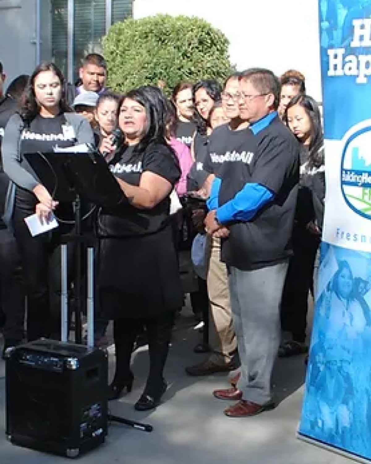 A woman at an outdoor podium speaking into a microphone, surrounded by supporters wearing matching black T-shirts.