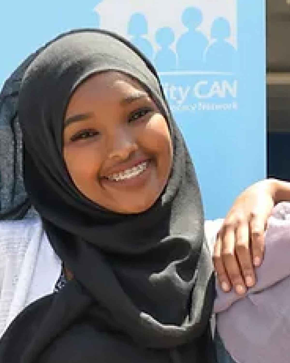 A smiling young person wearing a black hijab, posing in front of a bright blue banner.