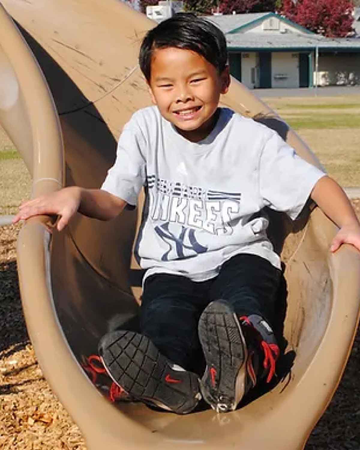 A smiling child wearing a gray T-shirt sliding down a tan plastic slide at a playground.