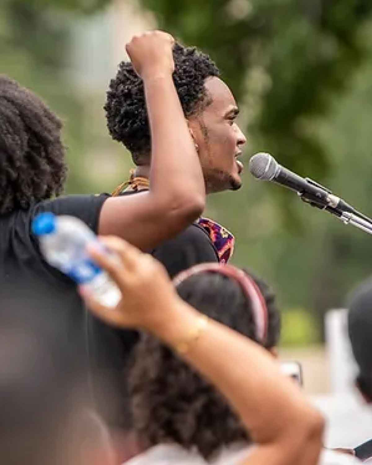 A man at an outdoor rally raising his fist in the air while speaking into a microphone, with onlookers in the foreground.