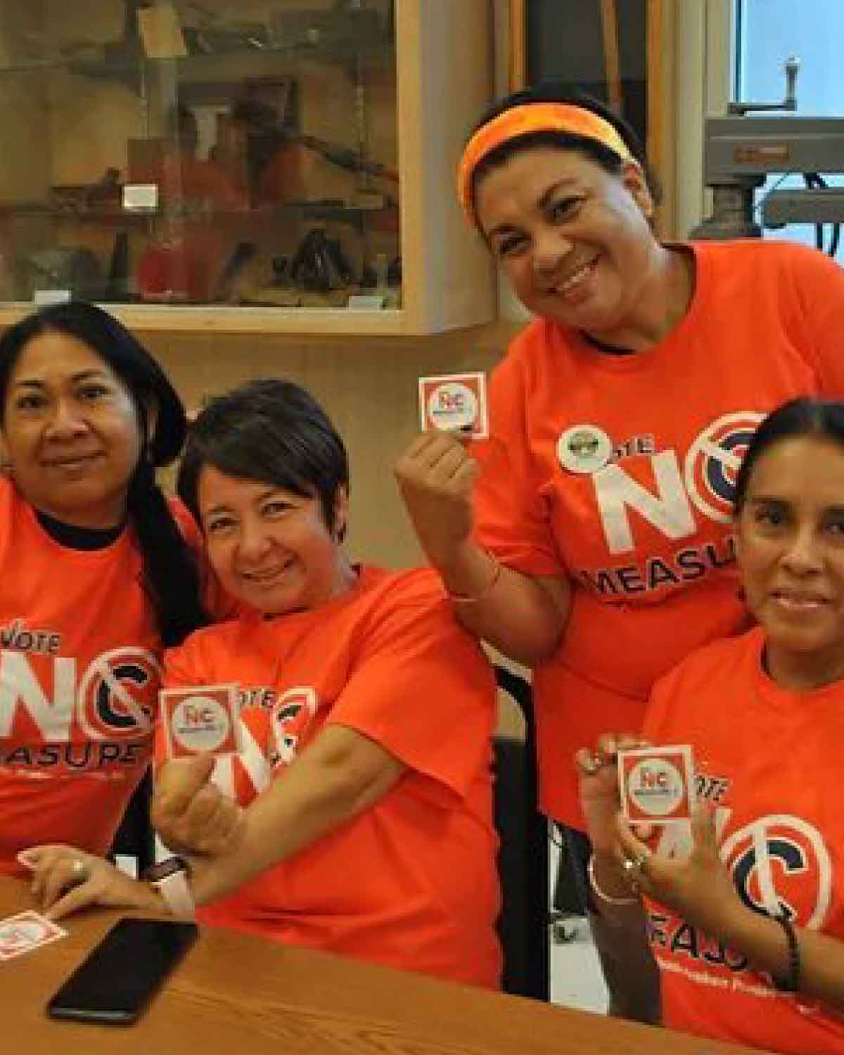 Four women wearing orange ‘No Measure C’ T-shirts seated together and holding up matching campaign stickers.