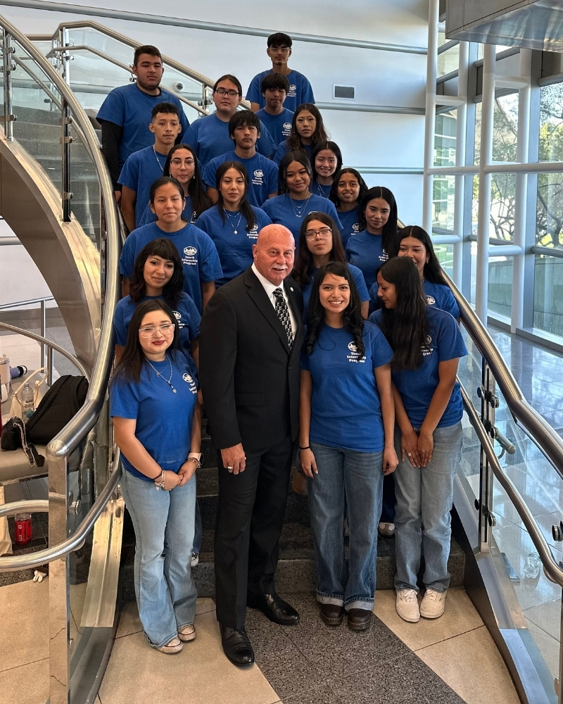 A group of students in matching blue shirts from Building Healthy Communities Fresno poses on a staircase with a Jerry Dyer at the front, inside a modern building with large glass windows.