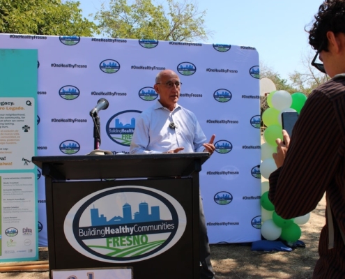 Community leader speaks at Calwa Park Renovation groundbreaking event hosted by Fresno Building Healthy Communities with #OneHealthyFresno signage in the background.