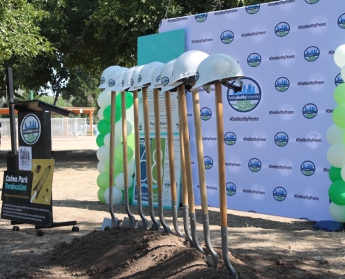 Line of shovels and hard hats ready for use at the Calwa Park groundbreaking, framed by trees and balloons.