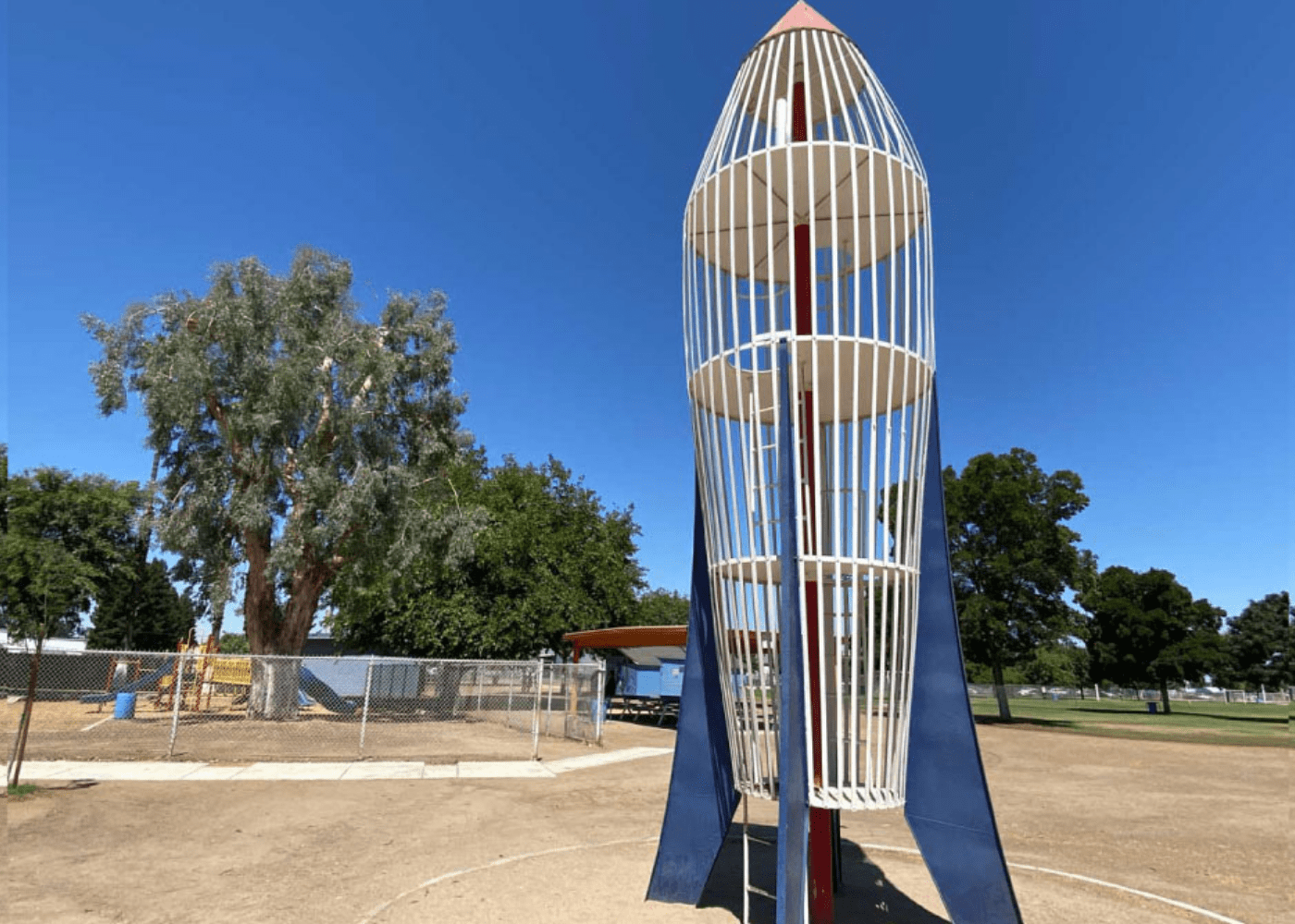 A tall, vintage rocket-shaped playground structure made of white metal bars and red accents stands in an open dirt area near a fenced playground and large shade trees under a clear blue sky.
