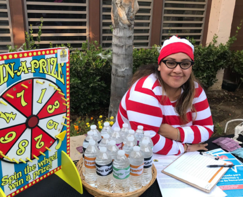 Volunteer dressed as Waldo hosting a Spin-A-Prize game booth at Fresno BHC’s Well-O-Ween