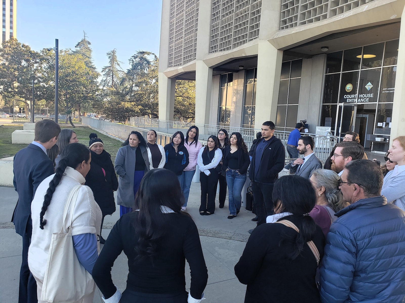 Did Caltrans Shortchange South Fresno? Judge Weighs Highway 99 Expansion Lawsuit A large group of people gathered in a loose circle on a concrete plaza outside a courthouse. They appear to be listening to a speaker, with the architectural details of the building's textured exterior visible in the background.