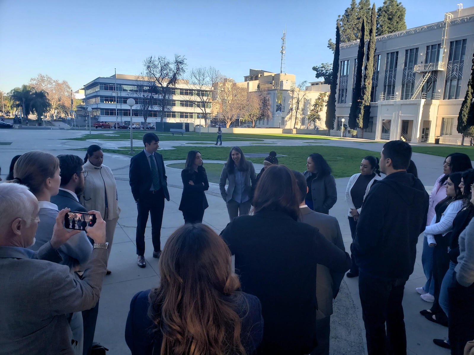 Did Caltrans Shortchange South Fresno? Judge Weighs Highway 99 Expansion Lawsuit A wide shot from behind a group of people standing in a circle on an outdoor plaza. In the background, there is a large green lawn, a white mid-century modern building, and tall cypress trees under a clear blue sky.