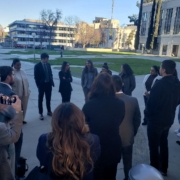 Did Caltrans Shortchange South Fresno? Judge Weighs Highway 99 Expansion Lawsuit A wide shot from behind a group of people standing in a circle on an outdoor plaza. One person on the left is holding up a smartphone to take a photo or video of the gathering. The background shows sun-drenched city buildings and a park.
