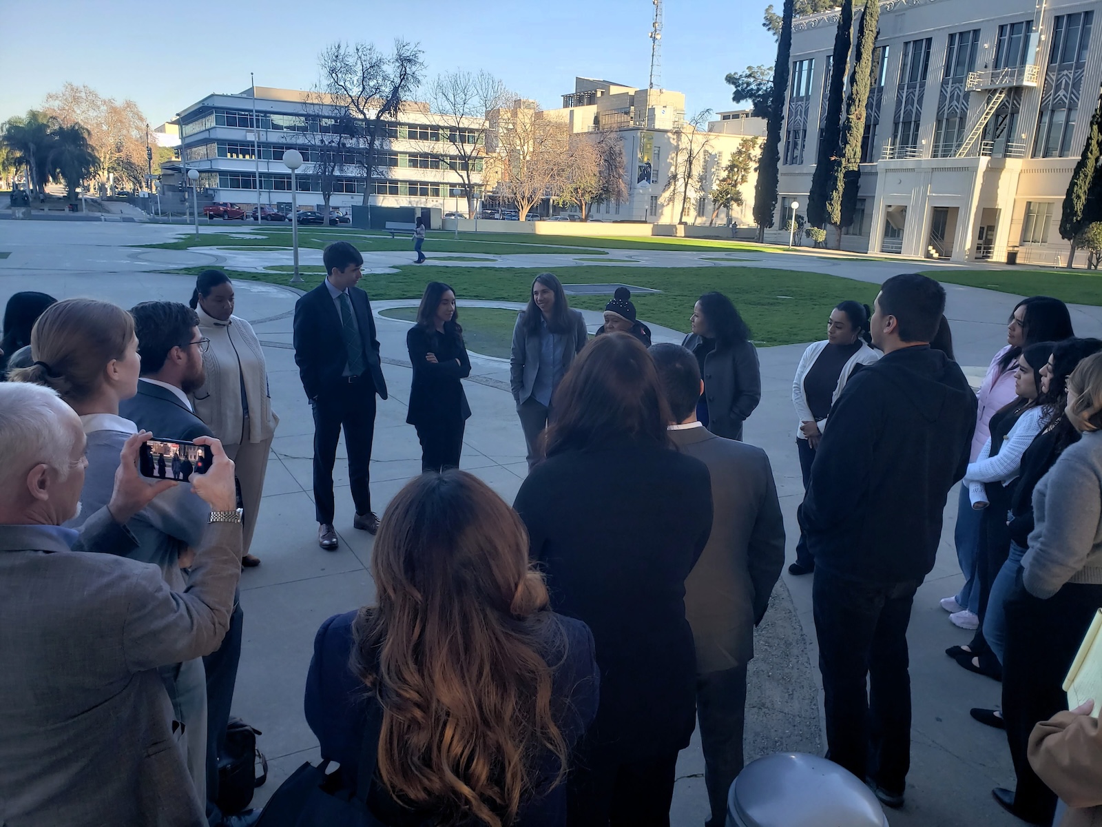 Did Caltrans Shortchange South Fresno? Judge Weighs Highway 99 Expansion Lawsuit A wide shot from behind a group of people standing in a circle on an outdoor plaza. One person on the left is holding up a smartphone to take a photo or video of the gathering. The background shows sun-drenched city buildings and a park.