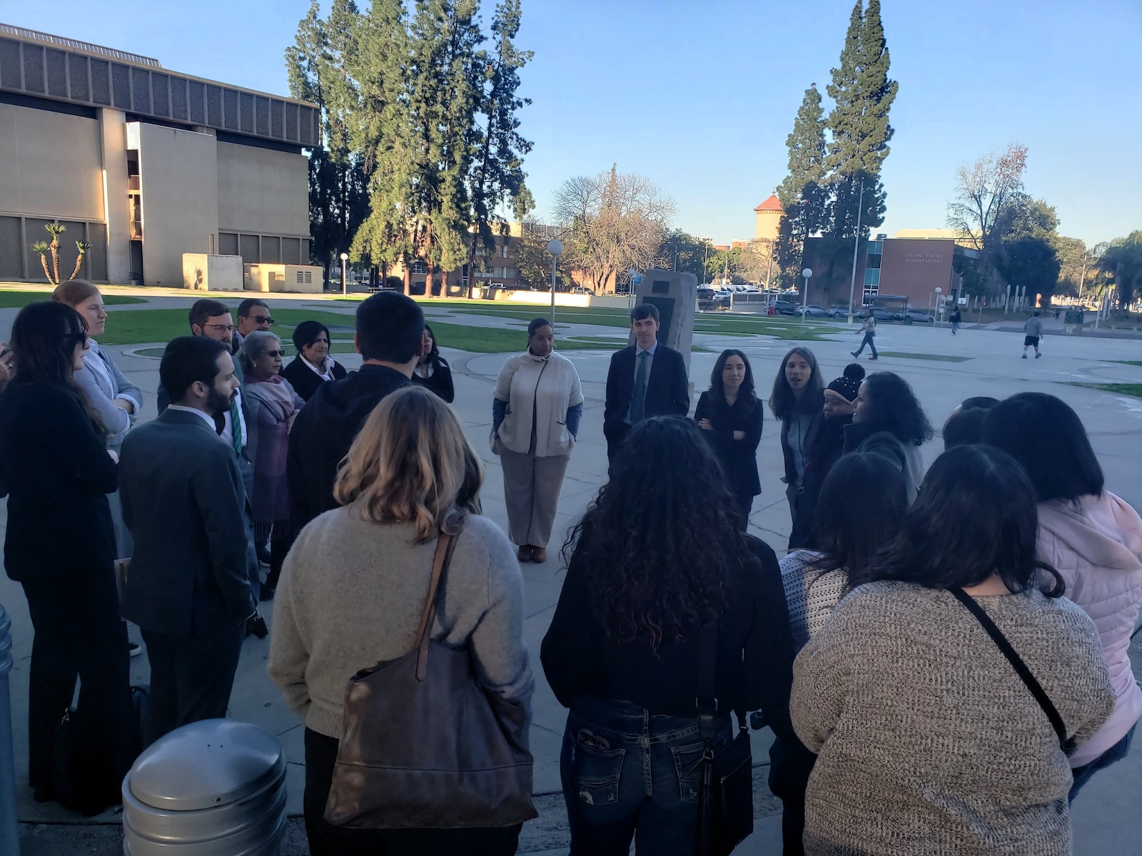 Did Caltrans Shortchange South Fresno? Judge Weighs Highway 99 Expansion Lawsuit A view from the opposite side of the circle, looking toward a different set of buildings and tall pine trees. The group is engaged in a discussion, with shadows stretching across the pavement in the afternoon light.