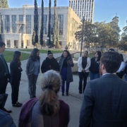 Did Caltrans Shortchange South Fresno? Judge Weighs Highway 99 Expansion Lawsuit A candid shot of the group discussion. A woman in a grey coat and blue trousers is speaking with her hands raised, while others in the circle listen attentively. The white courthouse building is visible in the background.