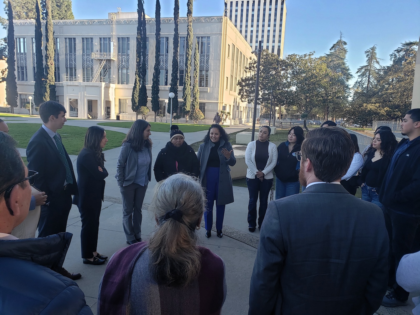 Did Caltrans Shortchange South Fresno? Judge Weighs Highway 99 Expansion Lawsuit A candid shot of the group discussion. A woman in a grey coat and blue trousers is speaking with her hands raised, while others in the circle listen attentively. The white courthouse building is visible in the background.