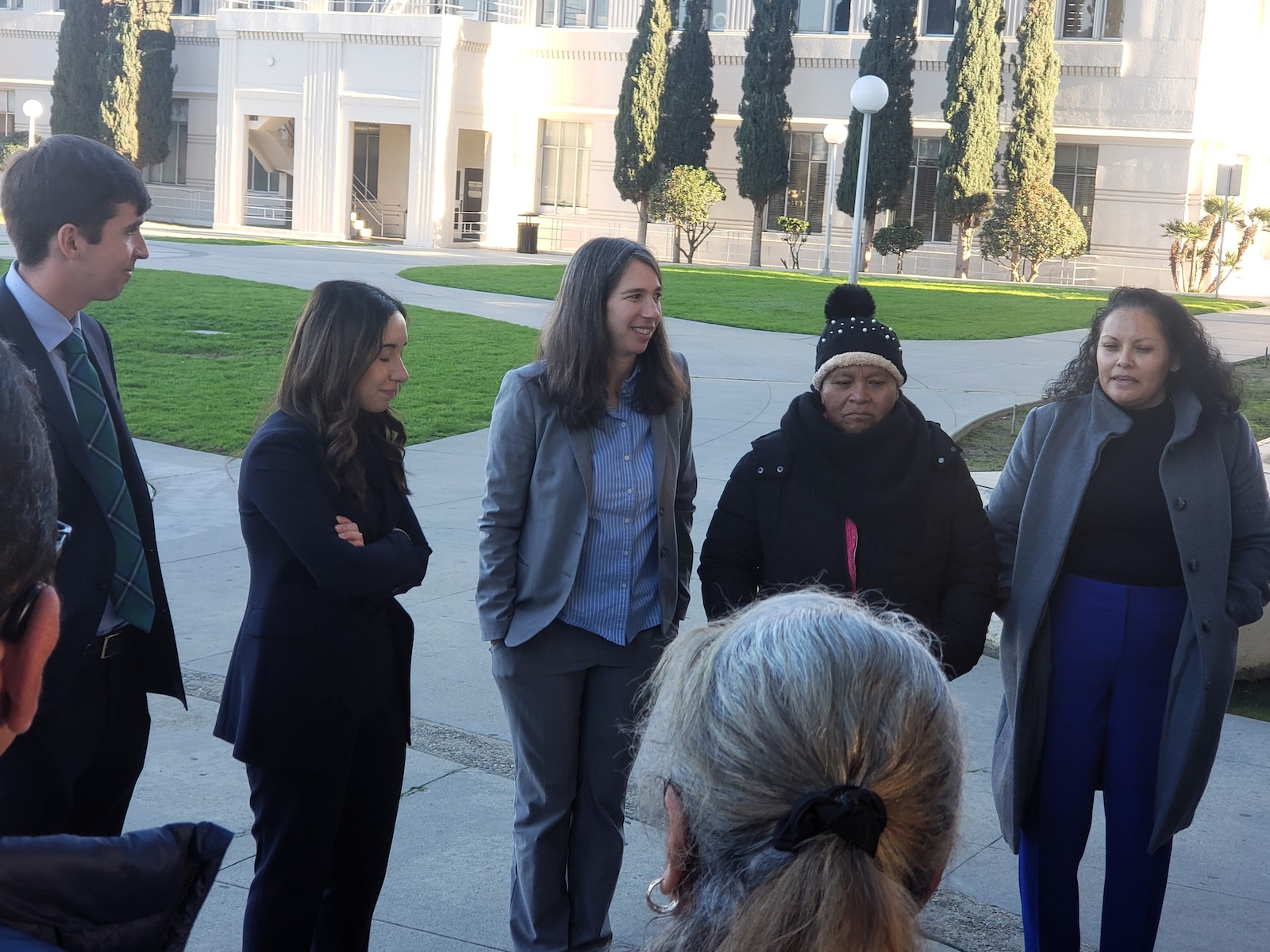 Did Caltrans Shortchange South Fresno? Judge Weighs Highway 99 Expansion Lawsuit A medium close-up of four people in the circle. A woman in a grey blazer is smiling and looking toward a woman in a dark coat and beanie. The lighting is bright and direct, highlighting their expressions during the conversation.