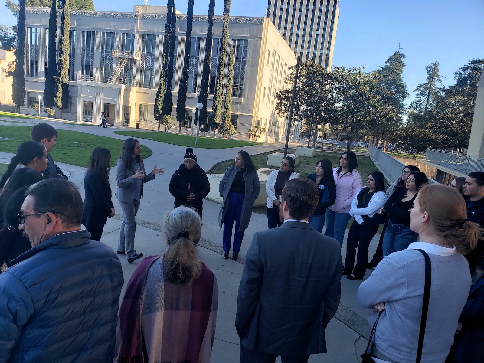 Did Caltrans Shortchange South Fresno? Judge Weighs Highway 99 Expansion Lawsuit An elevated wide shot of the group gathered on the plaza. A woman in a grey suit is gesturing while speaking to the crowd. The scene is framed by a mix of modern and classical architectural styles and mature trees.