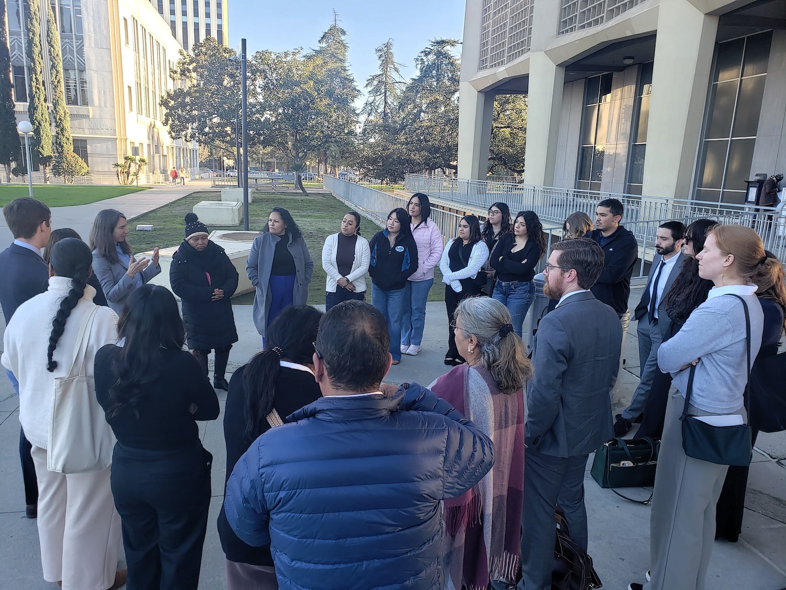 Did Caltrans Shortchange South Fresno? Judge Weighs Highway 99 Expansion Lawsuit A shot from behind the group, focusing on the speaker and the attentive audience. A man in a blue puffer jacket is in the foreground, and several people in professional suits are scattered throughout the circle.