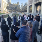 Did Caltrans Shortchange South Fresno? Judge Weighs Highway 99 Expansion Lawsuit A shot from behind the group, focusing on the speaker and the attentive audience. A man in a blue puffer jacket is in the foreground, and several people in professional suits are scattered throughout the circle.