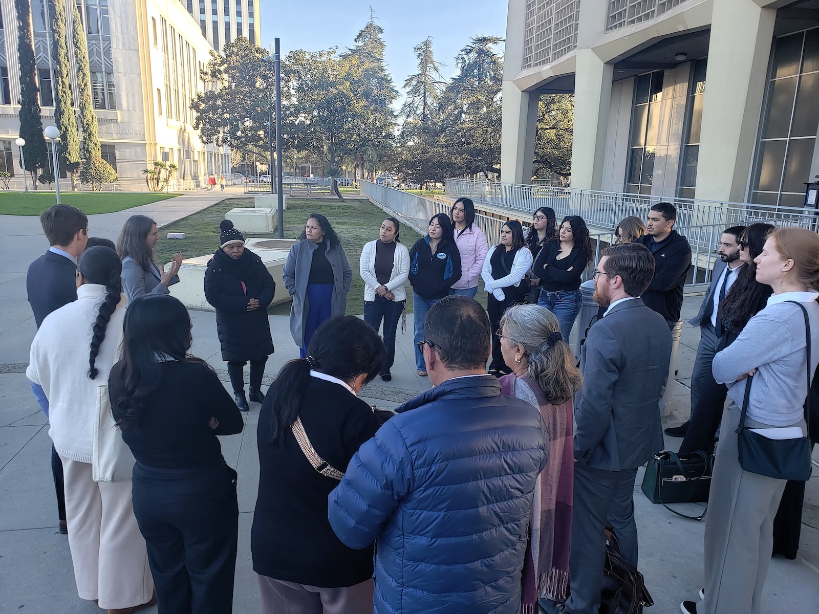Did Caltrans Shortchange South Fresno? Judge Weighs Highway 99 Expansion Lawsuit A shot from behind the group, focusing on the speaker and the attentive audience. A man in a blue puffer jacket is in the foreground, and several people in professional suits are scattered throughout the circle.