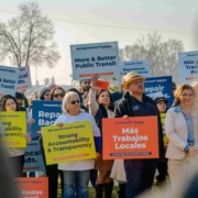 A close-up, candid shot of two men engaged in a friendly conversation during the event. One man wears a blue "Moving Fresno Forward" polo shirt, and the other is in a grey suit. In the blurred background, other attendees and the vibrant campaign balloon arch are visible.