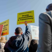 A low-angle shot looking up at supporters holding signs against a clear blue sky. A yellow sign reading "Strong Accountability & Transparency" is held high in the center. Other visible signs include "Create Local Jobs" and "Fix Local Roads," emphasizing the key themes of the gathering.