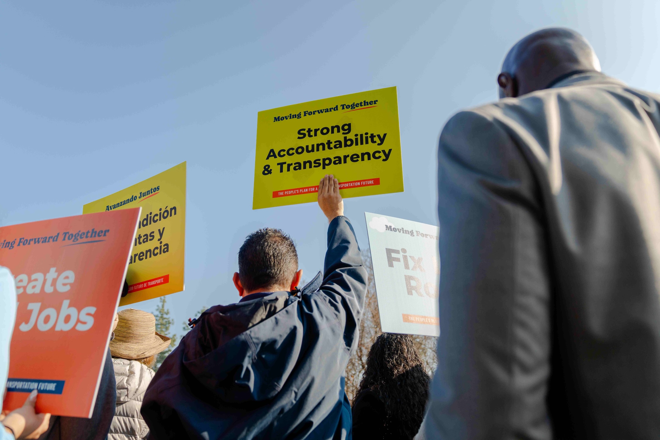 A low-angle shot looking up at supporters holding signs against a clear blue sky. A yellow sign reading "Strong Accountability & Transparency" is held high in the center. Other visible signs include "Create Local Jobs" and "Fix Local Roads," emphasizing the key themes of the gathering.