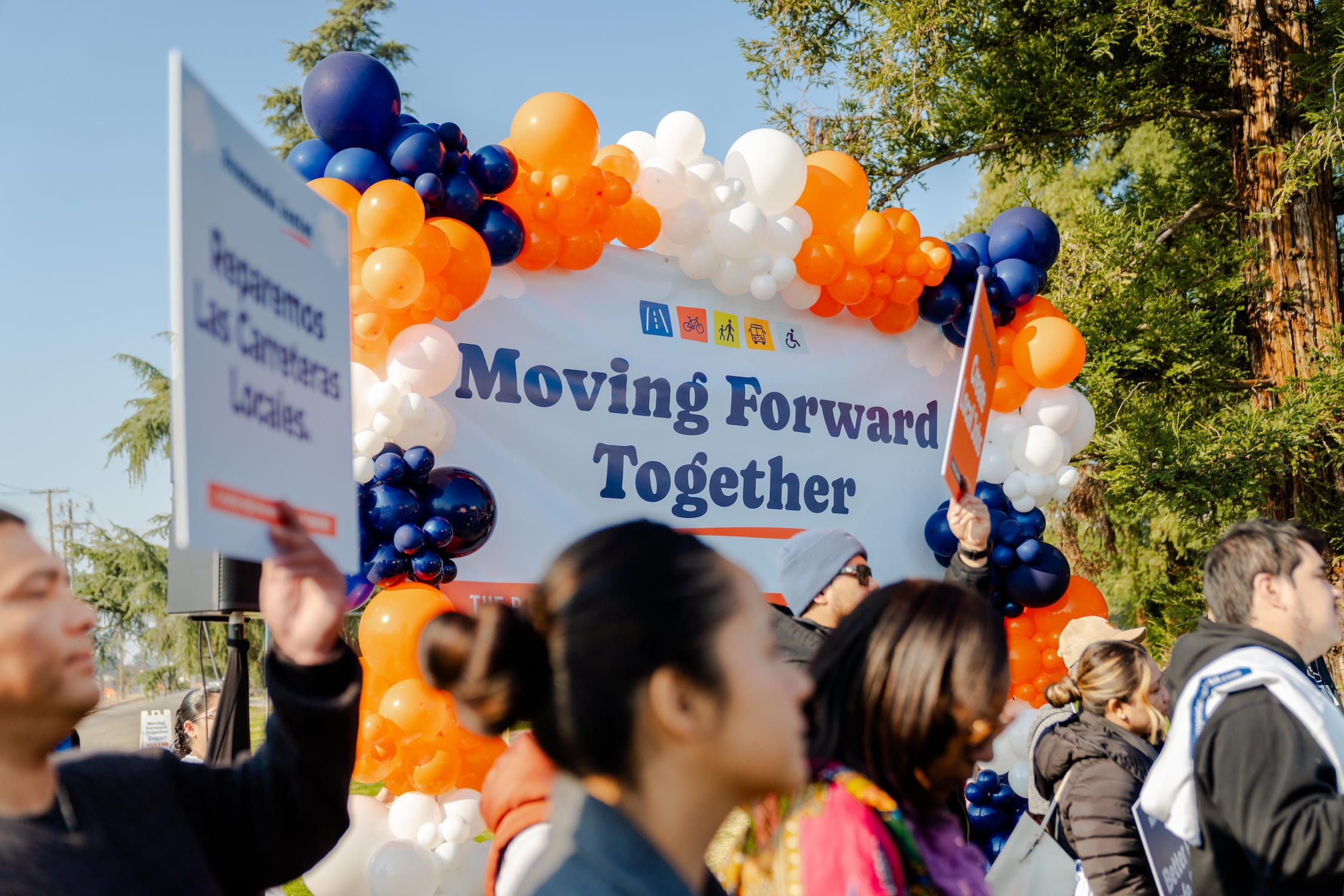 A group of approximately a dozen diverse community members and speakers stand together for a group photo behind the "Better Roads. Safe Streets." podium. They are holding a variety of signs, including "Create Local Jobs" and "Safe Routes to School," with a large orange and blue balloon arch behind them.