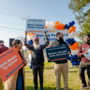 Four individuals standing on lawn, each holding colorful signs stating "Safe Routes To School," "More & Better Public Transit," "Better Roads. Safe Streets," and "Fix Local Roads."