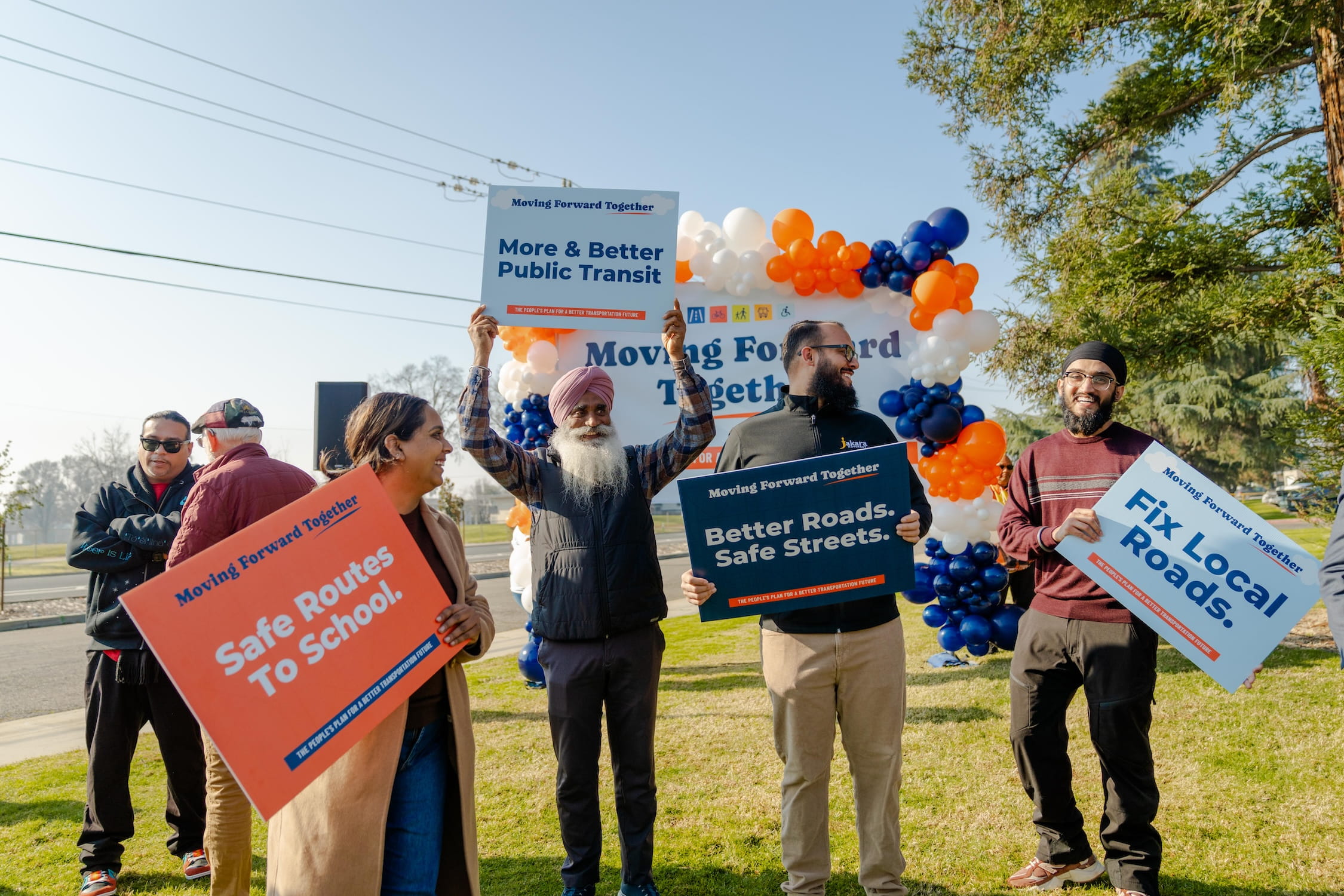 Four individuals standing on lawn, each holding colorful signs stating "Safe Routes To School," "More & Better Public Transit," "Better Roads. Safe Streets," and "Fix Local Roads."