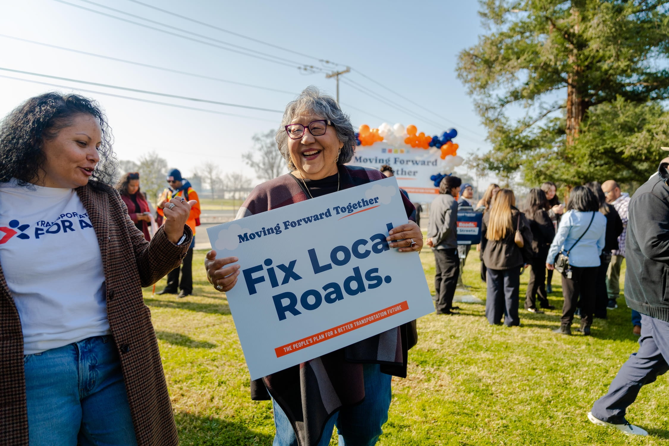 Two women standing together in conversation. One woman holding blue sign that reads, "Fix Local Roads."