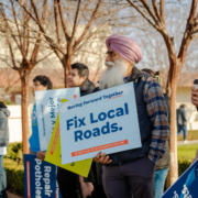 A row of supporters listening to the speaker. They are holding colorful signs, with one stating, "Fix Local Roads."